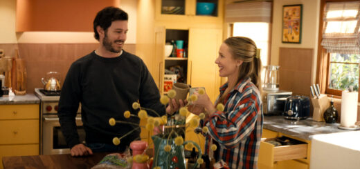"A man and woman smile and toast with mugs in a cozy, sunlit kitchen, surrounded by yellow cabinets, kitchen appliances, and decorative items, creating a warm, homey atmosphere."