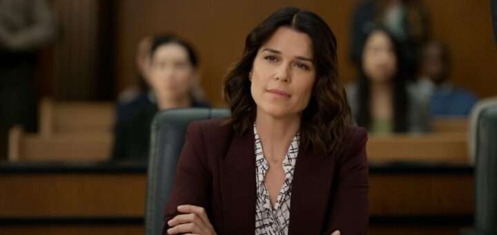 "Woman in a blazer sits with folded arms in a courtroom, looking thoughtful; blurred audience members are seated behind her, suggesting a legal proceeding or trial setting."