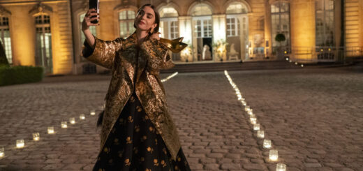 "Woman in a gold coat and black dress takes a selfie with her phone at night in a candlelit courtyard outside an elegant, well-lit building with large windows and cobblestone ground."