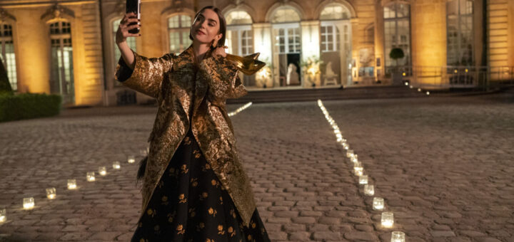 "Woman in a gold coat and black dress takes a selfie with her phone at night in a candlelit courtyard outside an elegant, well-lit building with large windows and cobblestone ground."