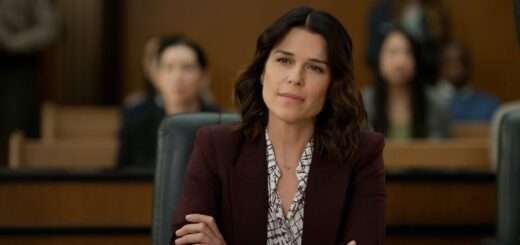 "Woman in a blazer sits with folded arms in a courtroom, looking thoughtful; blurred audience members are seated behind her, suggesting a legal proceeding or trial setting."